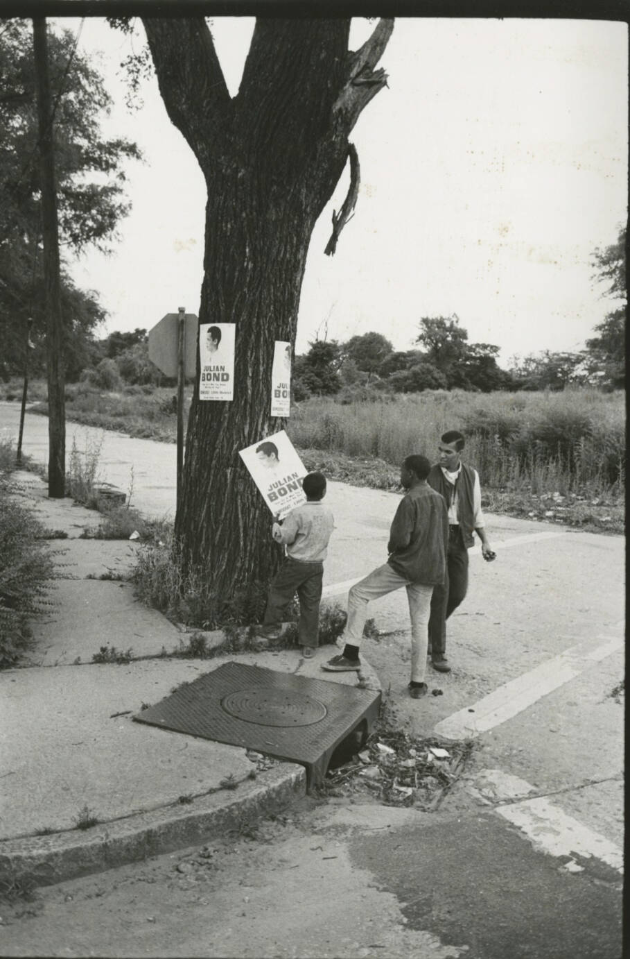Black and white photograph of two men and a child hanging up posters on the corner of a street with a tree.